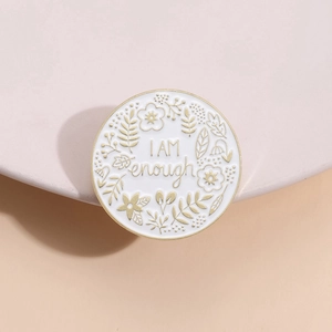 Round enamel 'I AM enough' pin with golden floral patterns on a ceramic display