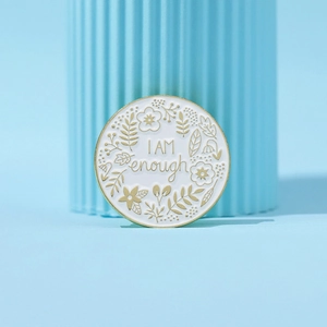 White enamel brooch with golden floral border and I AM ENOUGH text on blue stand