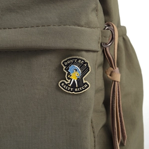 Detail der Emaille-Anstecknadel Mädchen im Regenmantel auf grüner Tasche mit Spruch