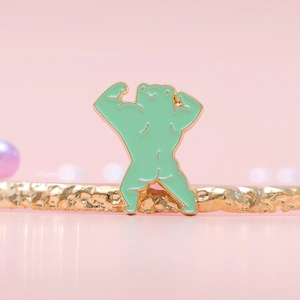 Funny green muscular frog enamel pin with gold metal edges on a pink display background