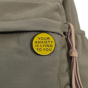 Pin's rond en émail jaune avec texte 'YOUR ANXIETY IS LYING TO YOU' sur un sac vert