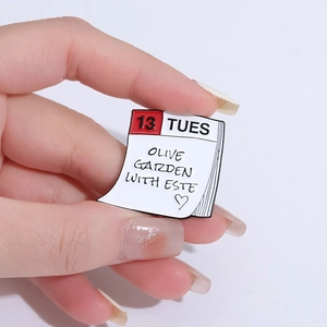 Handheld Olive Garden with Este calendar enamel pin featuring date 13 Tuesday and heart