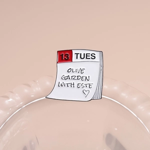 Calendar enamel pin featuring Olive Garden with Este and 13 Tuesday text on pink backdrop