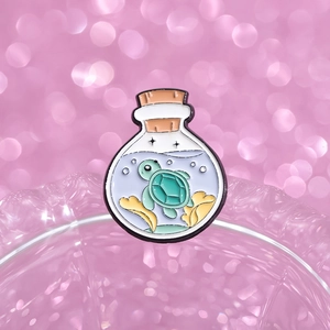 Enamel pin featuring a green turtle in a drift bottle with pink bokeh background