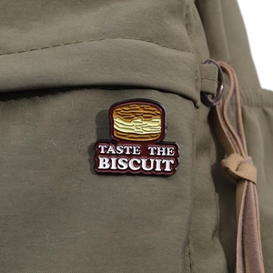Detailed close-up of a layered biscuit enamel pin with Taste The Biscuit text on canvas
