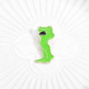 Funny green frog enamel pin with long legs displayed on a decorative white porcelain plate