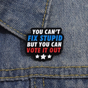 Metal lapel pin with text You Can't Fix Stupid But You Can Vote It Out on denim