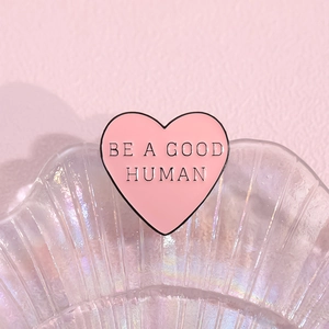 Minimalist pink heart enamel pin with Be A Good Human text on a shell texture tray