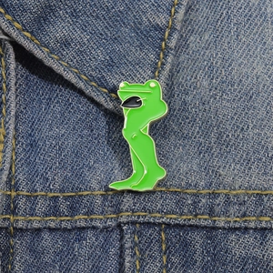 Funny cartoon green frog enamel pin with long legs and sunglasses on denim jacket
