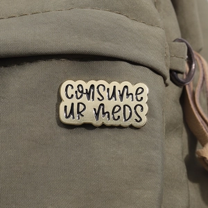 Detail of Consume Ur Meds metal enamel pin attached to a green canvas bag fabric