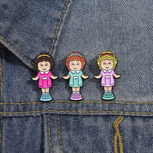 Three cute cartoon girl enamel pins with colorful dresses pinned on a blue denim jacket collar