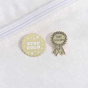 Golden enamel Stay Gold slogan medal and ribbon brooches on white canvas background