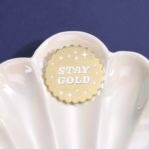 Spilla a medaglia dorata con scritta Stay Gold su vassoio a conchiglia bianca