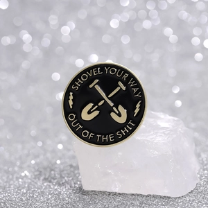 Black round enamel shovel pin with crossed shovels and motivational text on crystal