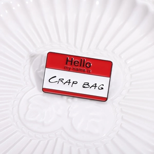 Red 'Hello my name is' enamel pin with 'CRAP BAG' text on a white textured plate