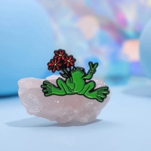 Cartoon green frog enamel pin holding red flowers with a V-sign on pink crystal stone