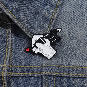 Detailed enamel pin featuring a hand holding a tattoo machine with a red heart on denim background