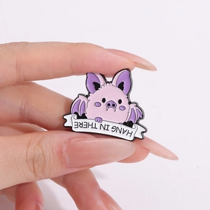 Cute pink cartoon bat enamel pin with Hang In There banner held by hand for scale