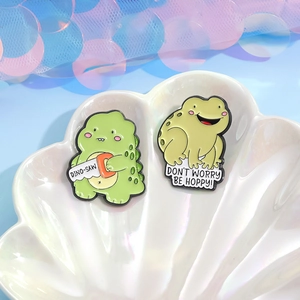 Cartoon enamel pin set with green dinosaur and frog on a white ceramic shell tray