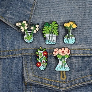 Fünf Emaille-Pflanzen-Pins in Laborflaschen auf einer blauen Jeansjacke dekoriert