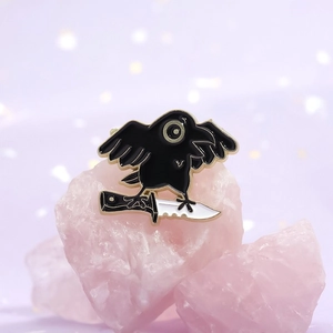 Detailed view of dark humor crow holding a knife enamel pin on pink crystal stone