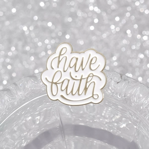 White enamel brooch with golden Have Faith script hanging on a glass rim with bokeh light