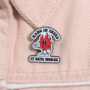 Macro shot of red enamel flame pin with 'Hang in there' and 'It gets worse' text on pink fabric