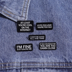 Detail of five black enamel slogan pins on blue denim jacket showing text and metal finish