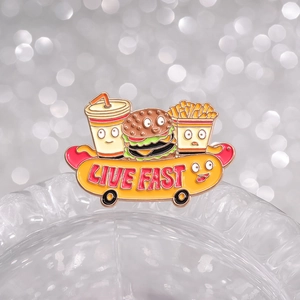 Cartoon fast food enamel pin with burger, fries and cola on a live fast hot dog skateboard