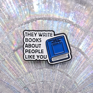 Blue DSM-5 book enamel pin with artistic text on a colorful iridescent background
