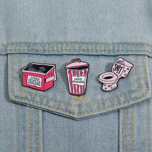 Dettaglio di tre spille in smalto rosa con scritte ironiche su denim
