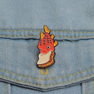 Detail of cute red fish enamel pin biting a sandwich on light blue denim fabric