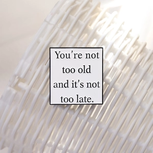 Square enamel pin badge with inspirational quote on white background with black metal edges