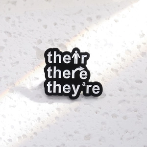Minimalist black enamel grammar brooch featuring their/there/they're educational text