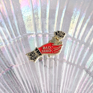 Unique hand holding black rose enamel pin with Bad Seed text and gold metal edges