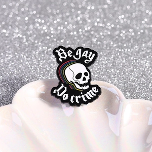 Skeleton enamel pin with rainbow hair and 'Be Gay Do Crime' text on a shell prop