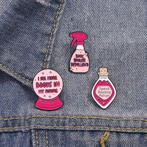 Three pink enamel pins with book themes styled on a blue denim jacket pocket