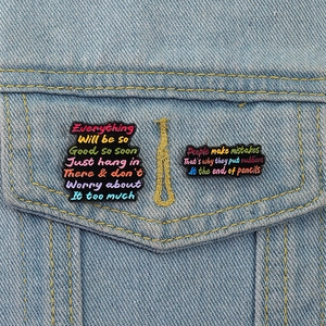 Close-up of colorful motivational enamel pins on a denim jacket pocket with text engraving