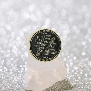 Round black enamel lapel pin with gold quote and decorative patterns on crystal base