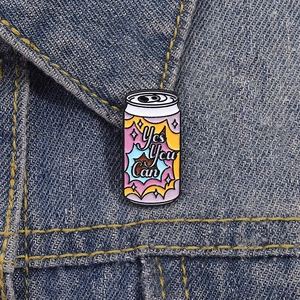 Macro shot of 'Yes You Can' soda can enamel pin attached to a blue denim jacket collar
