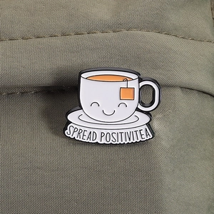 Close-up of white enamel cup pin with SPREAD POSITIVITEA text on green fabric