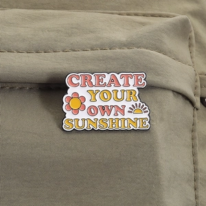 Dettaglio della spilla smaltata con scritta Create Your Own Sunshine su borsa in tessuto