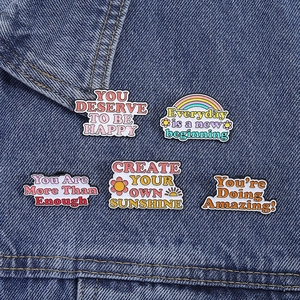 Dettaglio di cinque spille smaltate con citazioni motivazionali e arcobaleno su denim