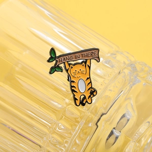 Detail der Emaille-Katzenbrosche am Zweig mit Text Hang In There auf gelbem Hintergrund