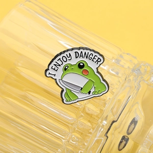 Detail of cartoon frog holding a knife enamel pin with 'I ENJOY DANGER' text