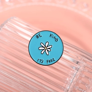 Makroaufnahme einer blauen Emaille-Brosche mit Gänseblümchen und BE KIND IT'S FREE-Slogan