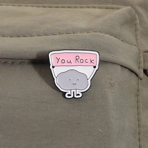 Macro shot of You Rock enamel pin on khaki canvas showing creative stone cartoon design
