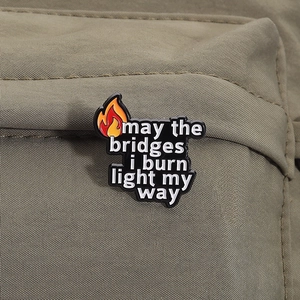 Emaille-Anstecknadel mit Flamme und Text: may the bridges i burn light my way am Rucksack