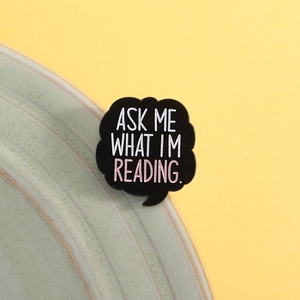 Gros plan du pin's 'Ask Me What I'm Reading' sur bord en céramique verte et fond jaune