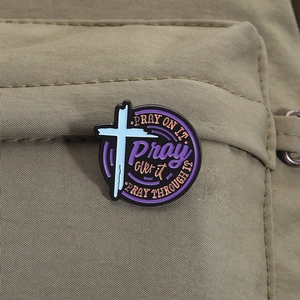 Detail of round purple enamel cross pin with prayer quote on a khaki canvas backpack